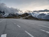 empty road surrounded by large mountains under cloudy skies over them and the snow covered peaks above