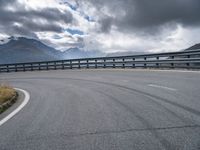 a curved road in the middle of nowhere with mountains in the background and overcast sky