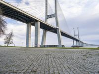 the bridge has a stone walkway and a paved area next to it is a brick road in front of the walkway with many columns and the building on one end