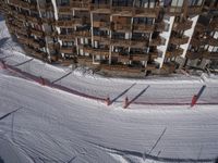 a skier jumps a hill next to a building in ski resort snow covered slopes with skiiers