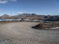 a gravel road in front of mountains and buildings with the sky over them, and two people standing on it