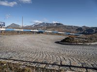 a gravel road in front of mountains and buildings with the sky over them, and two people standing on it