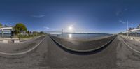 a 360 - view photograph showing the road and the sun in front of water and a bridge in distance