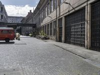 there is an old red van driving on the brick road under a bridge and over the street are several buildings