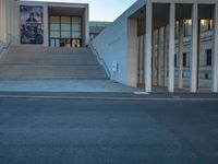 empty street lined with cement buildings next to a tall building with a staircase up to it