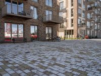 bikes parked outside an apartment complex with buildings in the background with a brick courtyard with two people in red jackets standing outside of it