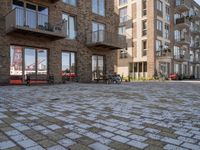 bikes parked outside an apartment complex with buildings in the background with a brick courtyard with two people in red jackets standing outside of it