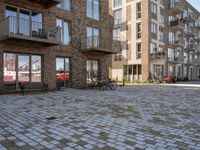 bikes parked outside an apartment complex with buildings in the background with a brick courtyard with two people in red jackets standing outside of it