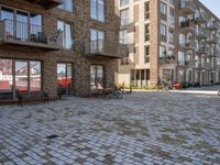 bikes parked outside an apartment complex with buildings in the background with a brick courtyard with two people in red jackets standing outside of it