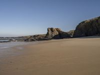 an empty beach with sand and rocks in the background with clear blue skies and water