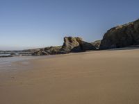 an empty beach with sand and rocks in the background with clear blue skies and water