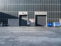 two garbage bins are sitting in a concrete parking lot, with the building's glass doors closed