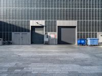 two garbage bins are sitting in a concrete parking lot, with the building's glass doors closed