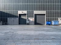 two garbage bins are sitting in a concrete parking lot, with the building's glass doors closed