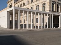 white building with columns at the entrance and people standing on the sidewalk outside it,