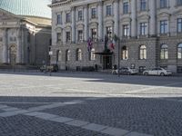 the european building features a paved stone courtyard and several old style cars parked in front