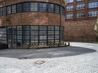 a brick circular building with windows and two benches in front of it on a brick pavemented area