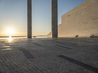 the sun set behind three large pillars and water with benches on it near the ocean