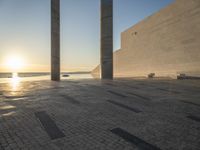 the sun set behind three large pillars and water with benches on it near the ocean