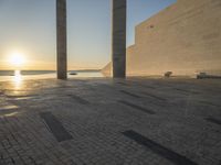 the sun set behind three large pillars and water with benches on it near the ocean