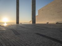 the sun set behind three large pillars and water with benches on it near the ocean