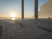 the sun set behind three large pillars and water with benches on it near the ocean