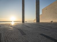 the sun set behind three large pillars and water with benches on it near the ocean