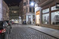 this is an empty street at night with a cobble stone sidewalk and parked cars