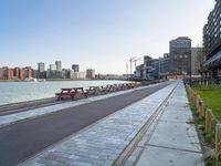 a paved path on a river side by the city and two empty benches lined up against a line