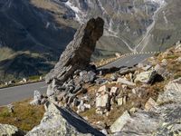 the winding mountain road is in between rocks and mountainside vegetations and a large rock formation in the foreground