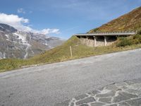 there is an empty asphalt road on the side of a mountain with snow on the mountains and green hills