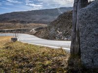 an asphalted road next to a mountain with a big rock at the side with grass, rocks and bushes and tall mountains behind