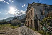 a house with the sun in the background sitting on a cobblestone road with an old building in front