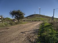 a dirt road near a mountain with wind turbines in the background and grassy area and plants