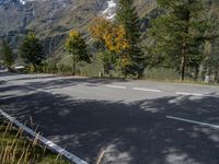 an empty road on the side of a mountain with trees on either side of it, and snow capped peaks in the background