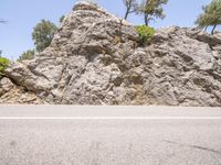 two people riding bicycles down the road past a rock wall and trees in the background