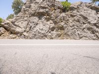 two people riding bicycles down the road past a rock wall and trees in the background