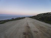 dirt road leads to the ocean by some grassy hills on shore at sunset of large cliff,