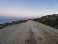 dirt road leads to the ocean by some grassy hills on shore at sunset of large cliff,