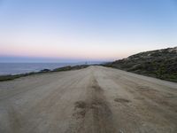 dirt road leads to the ocean by some grassy hills on shore at sunset of large cliff,