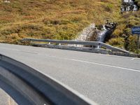 a highway in the middle of the mountains with a waterfall coming down it, beside a winding road