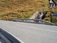 a highway in the middle of the mountains with a waterfall coming down it, beside a winding road