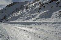 a snowy slope and a road with many snow covered tracks on it and one person on skis is snowboarding on the slope, with skis