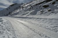 a snowy slope and a road with many snow covered tracks on it and one person on skis is snowboarding on the slope, with skis