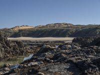 the view looking up at a beach and hill from a rocky shore line with the water on it