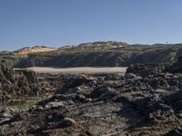 the view looking up at a beach and hill from a rocky shore line with the water on it