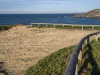 wooden fence and dirt path leading to beach by sea with blue sky above with mountains in background