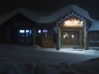 the entrance to a small building at night with snow covering the ground around it and a snow drifting onto the ground underneath