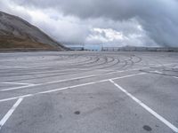 a car driving around a curve in an empty lot on a cloudy day with large mountains in the background