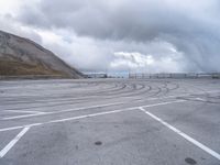 a car driving around a curve in an empty lot on a cloudy day with large mountains in the background